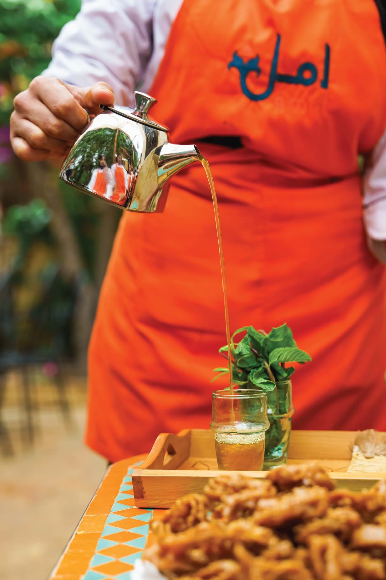An Amal trainee in an orange apron pouring Moroccan mint tea over a tray of pastries